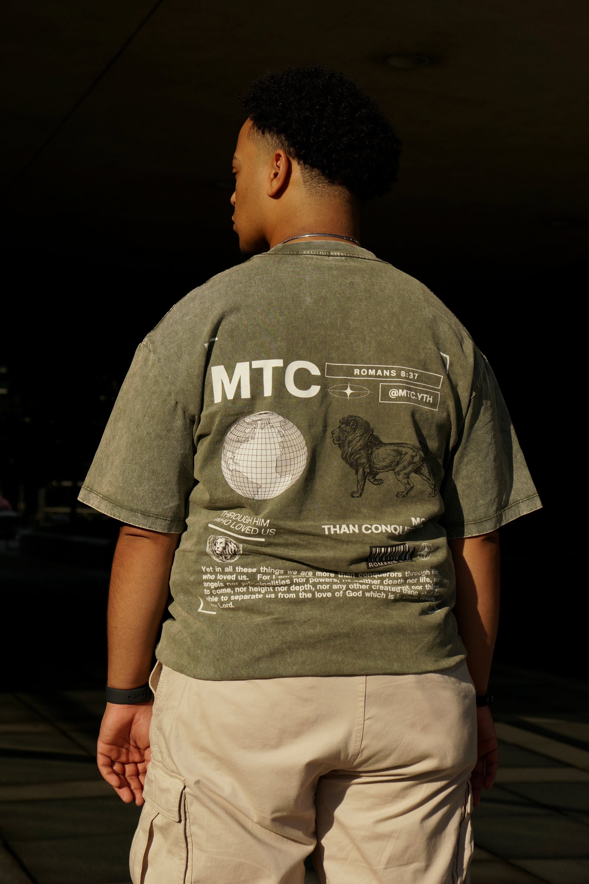 hype4holy christian apparel mtc streetwear oversized faded green t shirt, model picture, back | Person wearing a green t-shirt with 'MTC' branding and text, standing against a dark background.