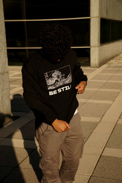hype4holy christian apparel oversized sweatshirt black color with be still white design model picture 1 | Person wearing a black sweater with a graphic christian design and text, standing in front of a modern building.