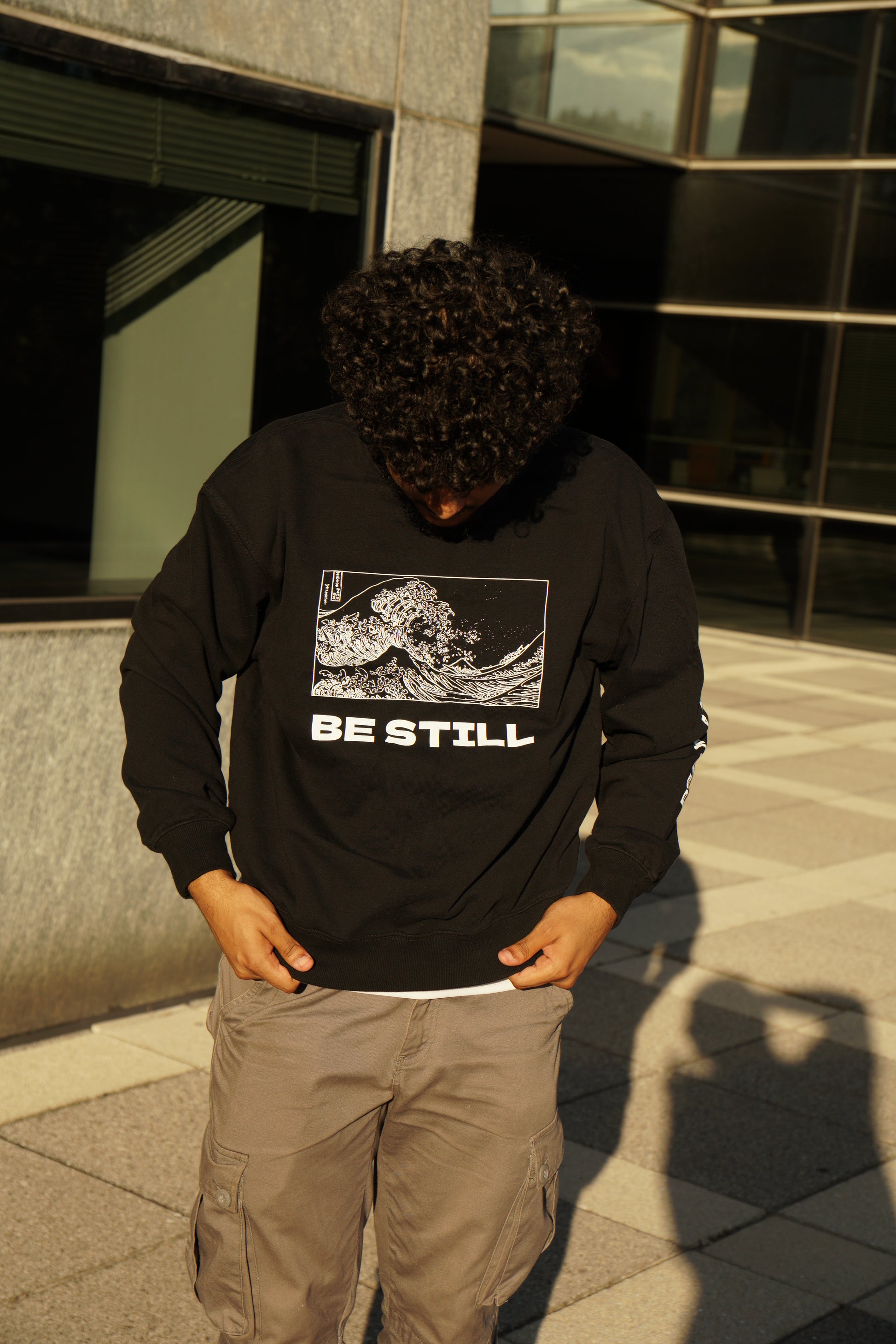 hype4holy christian apparel oversized sweatshirt black color with be still white design model picture 2 | Person wearing a black sweater with a graphic christian design and text, standing in front of a modern building.