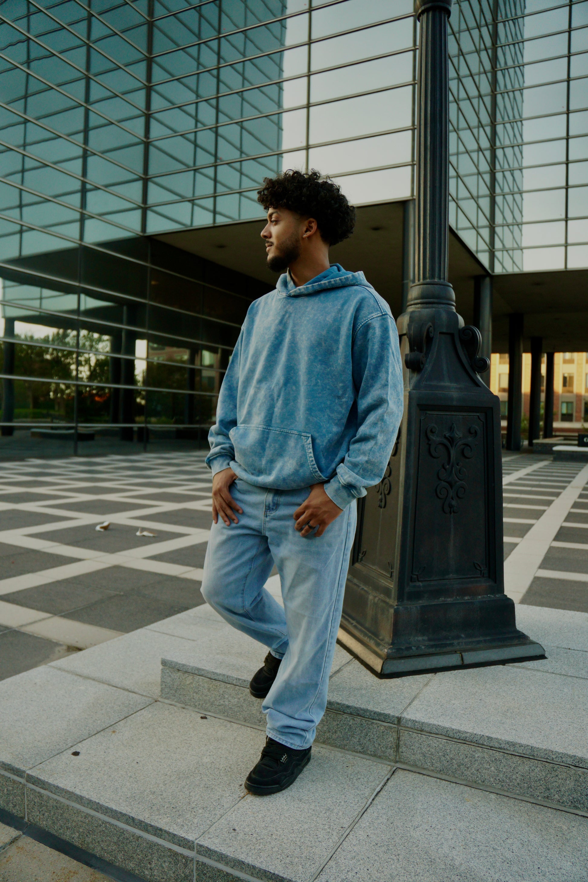 hype4holy christian apparel streetwear oversized faded hoodie blue color model picture front | Man in a blue hoodie and light blue jeans standing on steps in front of a modern building.