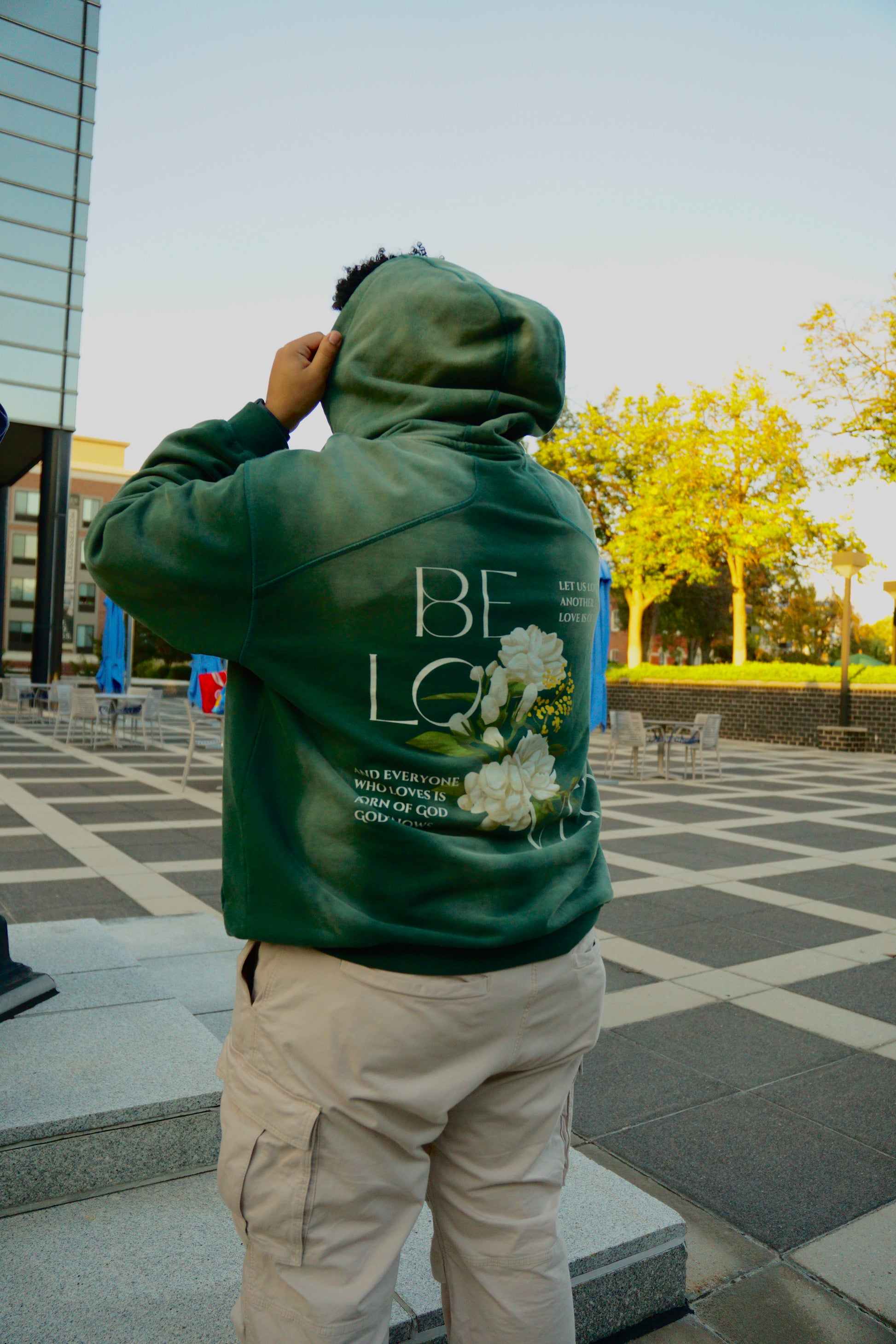 hype4holy christian apparel streetwear oversized heavyweight sun faded green hoodie with beloved flower design model picture standing, back |  Person wearing a green hoodie with floral design and text, standing outdoors.