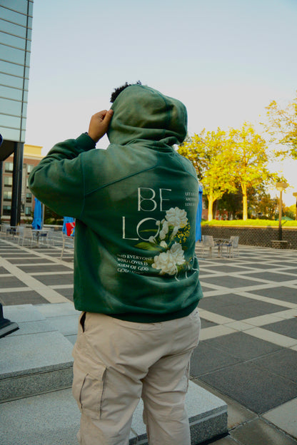 hype4holy christian apparel streetwear oversized heavyweight sun faded green hoodie with beloved flower design model picture standing, back |  Person wearing a green hoodie with floral design and text, standing outdoors.