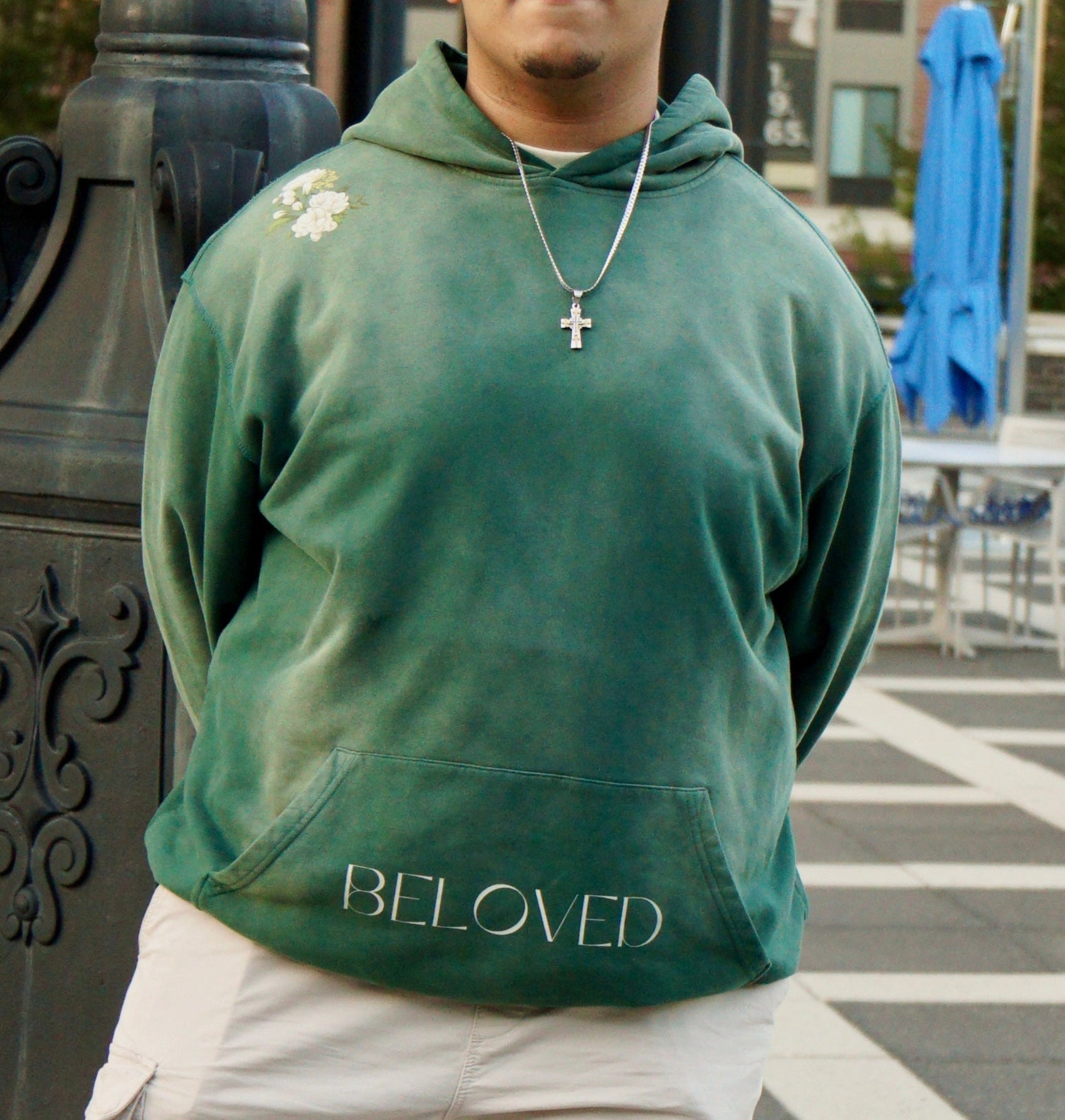hype4holy christian apparel streetwear oversized heavyweight sun faded green hoodie with beloved flower design model picture standing, back |  Person wearing a green hoodie with 'BELOVED' text, standing outdoors.