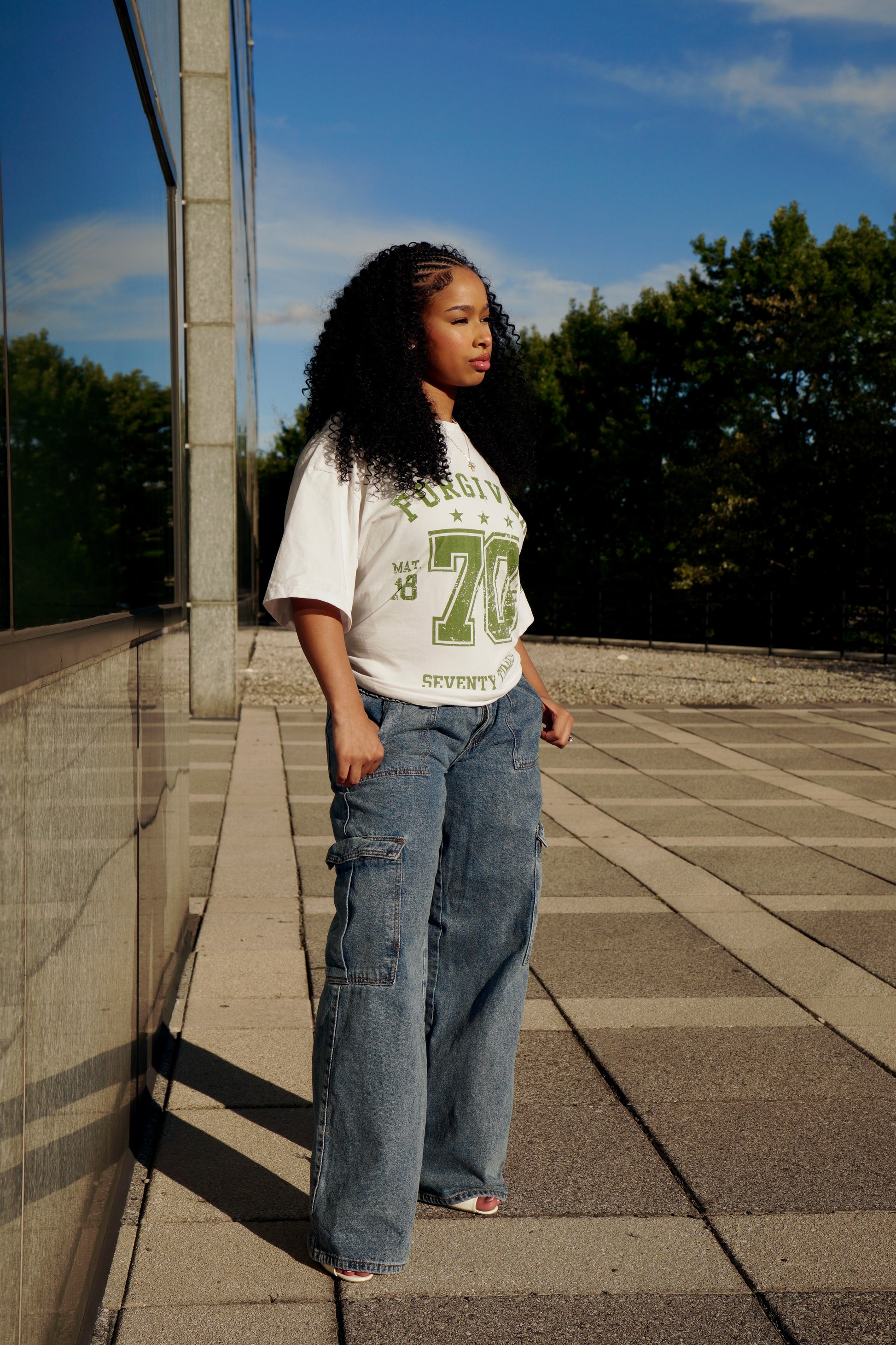 hype4holy christian streetwear unisex t-shirt white color forgiven design model picture profile | Person wearing a white t-shirt with green text and blue jeans standing on a paved area with trees in the background.