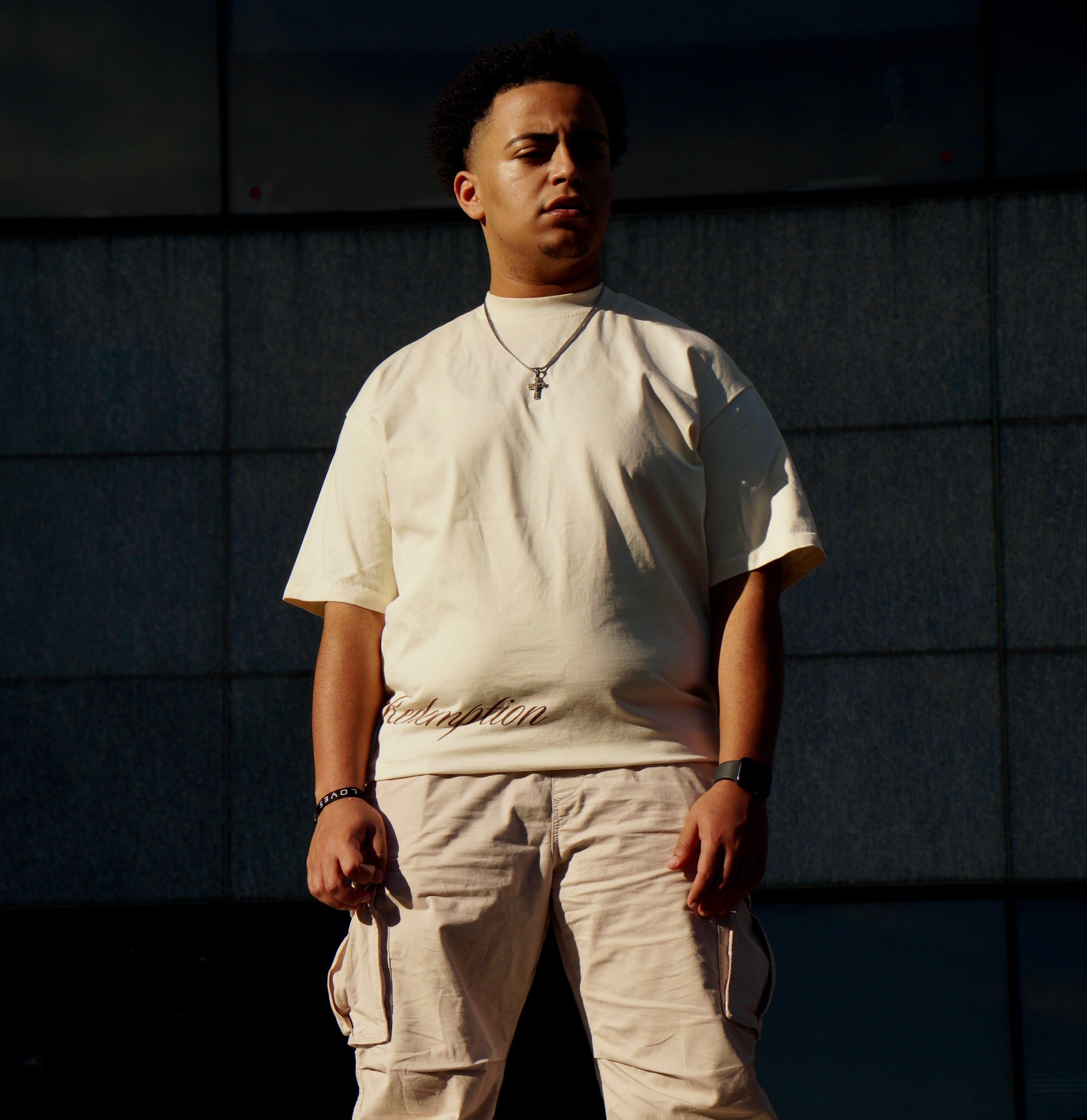 hype4holy christian streetwear t shirt model picture with redemption design in beige color in the front and beige pants standing on steps with a dark wall in the background